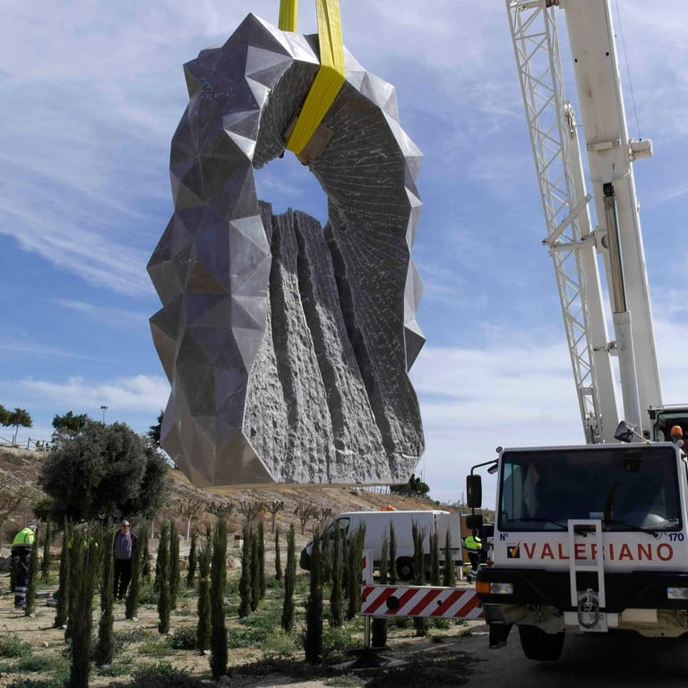 A white Valeriano industrial crane lifting a large geometric grey rock sculpture in a park.