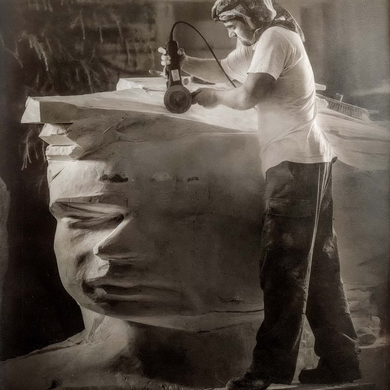 Artist using a power tool to carve a large stone head sculpture in a workshop.