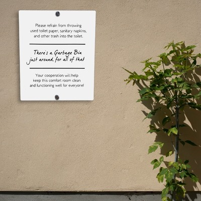 White acrylic sign with restroom instructions mounted on a beige wall next to a plant.