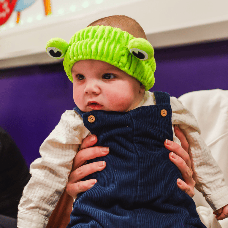 a baby wearing a a frog headband enjoying moo music class with her mum