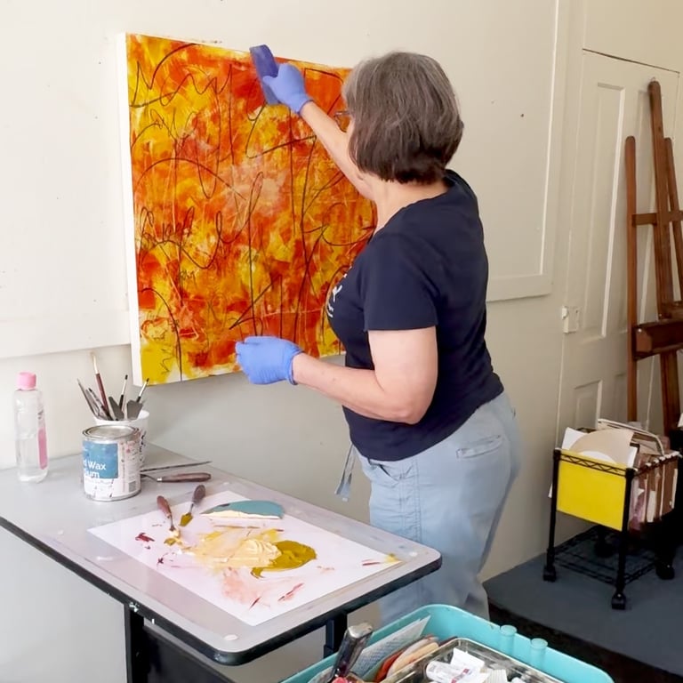 Artist in blue gloves applying cold wax medium to a large orange and yellow abstract painting.