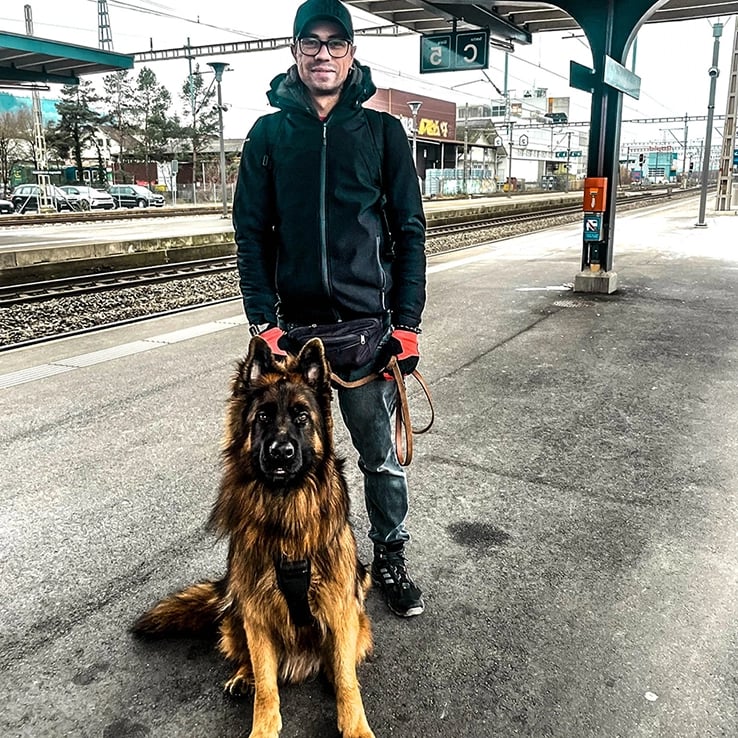 Foto eines lächelnden Mannes, der mit einem jungen Deutschen Schäferhund am Bahnhof steht