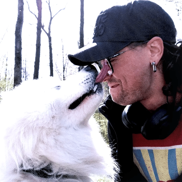 a photo of a man wearing a black baseball cap and glasses being licked in the face by white dog