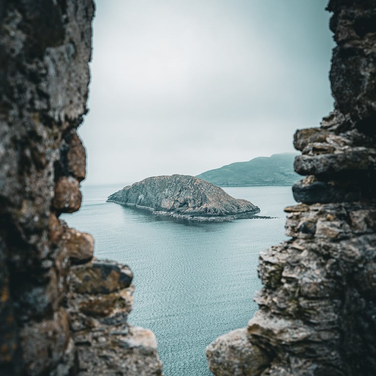 Vue d'une île rocheuse à travers les ruines du château de Duntulm, entourée d'une mer calme sous un ciel gris.