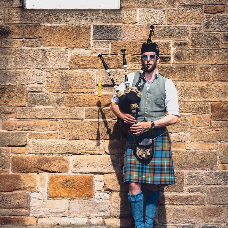 Joueur de cornemuse en kilt écossais, appuyé contre un mur de pierre à Édimbourg, baigné par la lumière du soleil.