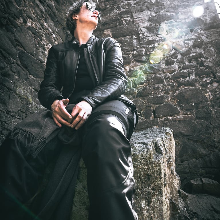 Personne en cuir noir, lunettes de soleil, assise dans des ruines. Lumière douce, posture contemplative, pierres texturées.
