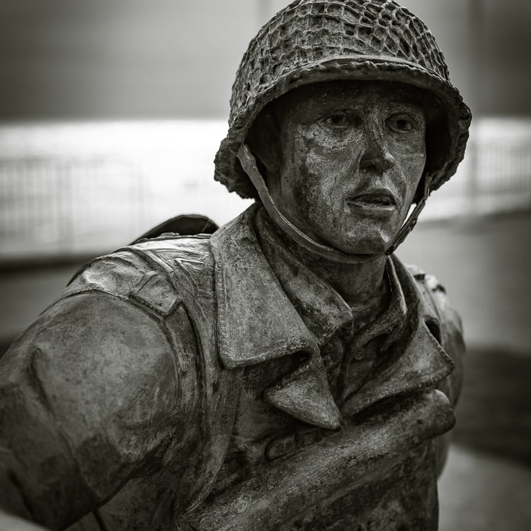 Gros plan sur le visage d'une statue de soldat en bronze, casque sur la tête, regard intense. Mer et ciel couvert en fond.