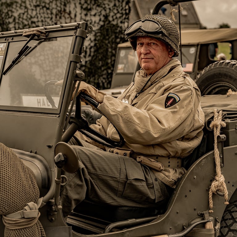 omme en uniforme de la Seconde Guerre assis dans une jeep vintage, un autre véhicule militaire visible en arrière-plan.