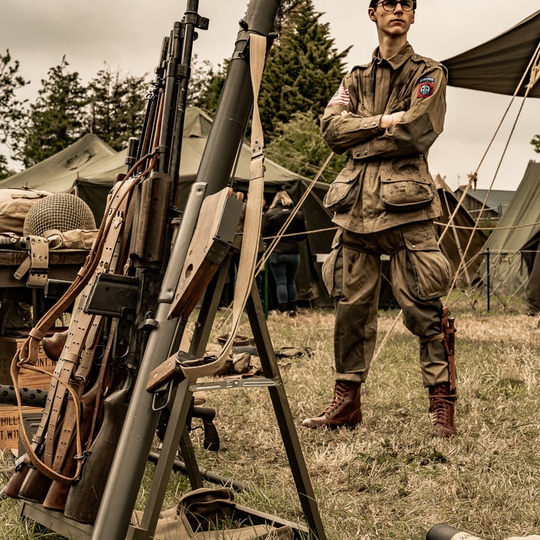 Soldat parachutiste dans un camp, bras croisés et regard au loin. Armes alignées sous un ciel sombre, tension palpable.