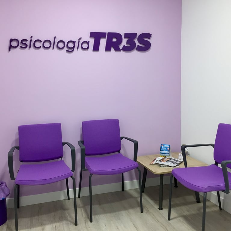 Sala de espera con tres sillas moradas, una mesa baja y una papelera. Se ve el logo de psicologíaTR3S en la pared