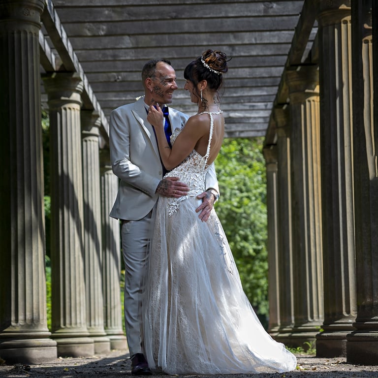 Ein Hochzeitspaar blickt sich verliebt an, während es unter einer Holzpergola mit Steinsäulen steht; die Braut trägt ein träg