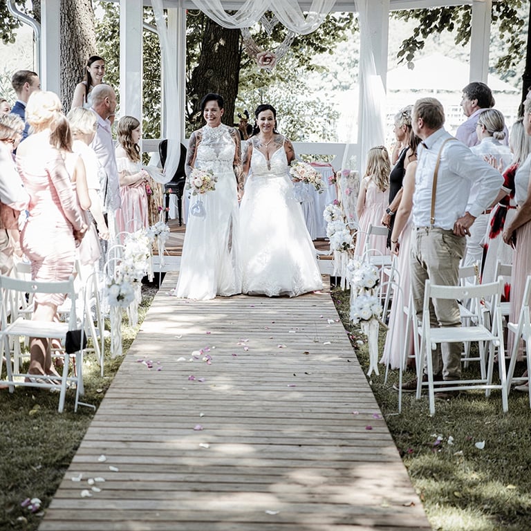 Dieses Farbfoto zeigt zwei Bräute, die in ihren Hochzeitskleidern einen Holzsteg entlanggehen, der mit verstreuten Blütenblät