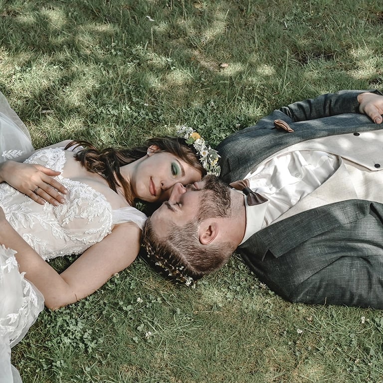Dieses Farbfoto zeigt ein Brautpaar, das auf grünem Gras liegt und sich ansieht. Die Braut trägt ein weißes Hochzeitskleid mi