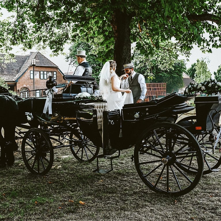 Dieses Farbfoto zeigt eine Hochzeitskutsche mit Pferden und drei Personen unter großen, belaubten Bäumen. Eine Braut in einem
