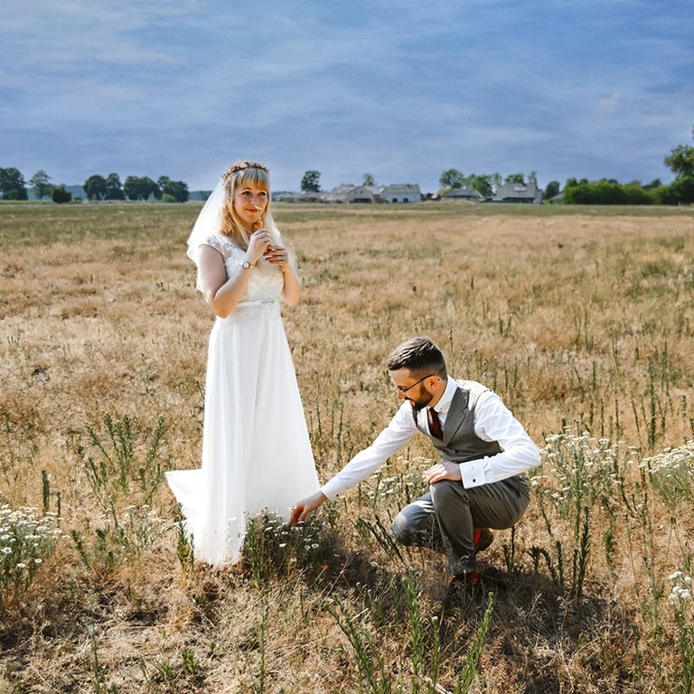  Dieses Farbfoto zeigt ein Brautpaar in einem trockenen Feld unter einem dramatischen Himmel. Die Braut steht in einem weißen