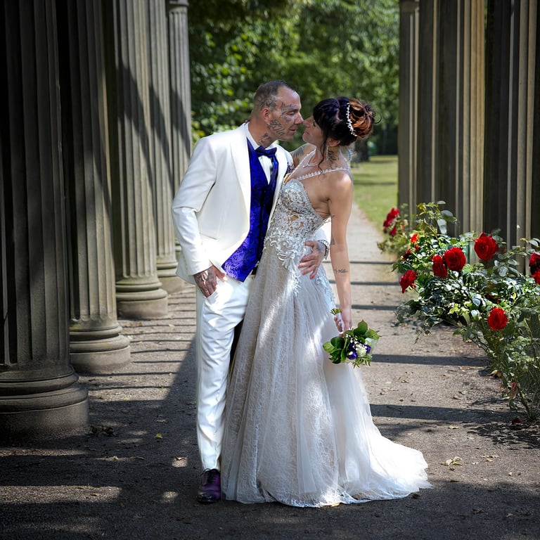 Ein Hochzeitspaar küsst sich unter einer Pergola mit Steinsäulen und einer Reihe roter Rosen; der Bräutigam trägt einen weiße