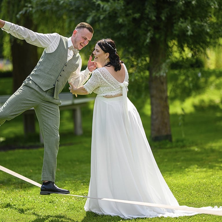 Dieses Farbfoto zeigt ein Brautpaar in einer spielerischen Pose auf einer Slackline in einem grünen Park. Der Bräutigam, gekl