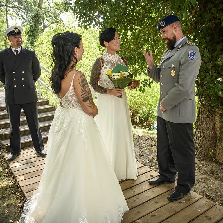 wei Bräute und zwei Männer in Militäruniformen unter einem Baum auf einem Holzsteg, eine Braut hält einen Blumenstrauß.