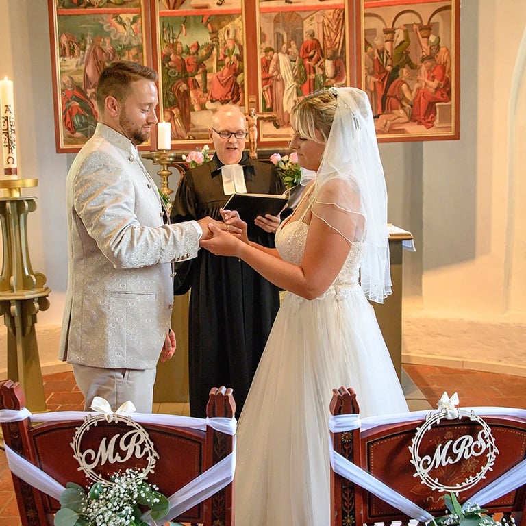 Dieses Farbfoto zeigt eine Hochzeitszeremonie in einer Kirche. Im Mittelpunkt stehen Braut und Bräutigam, die sich gegenseiti