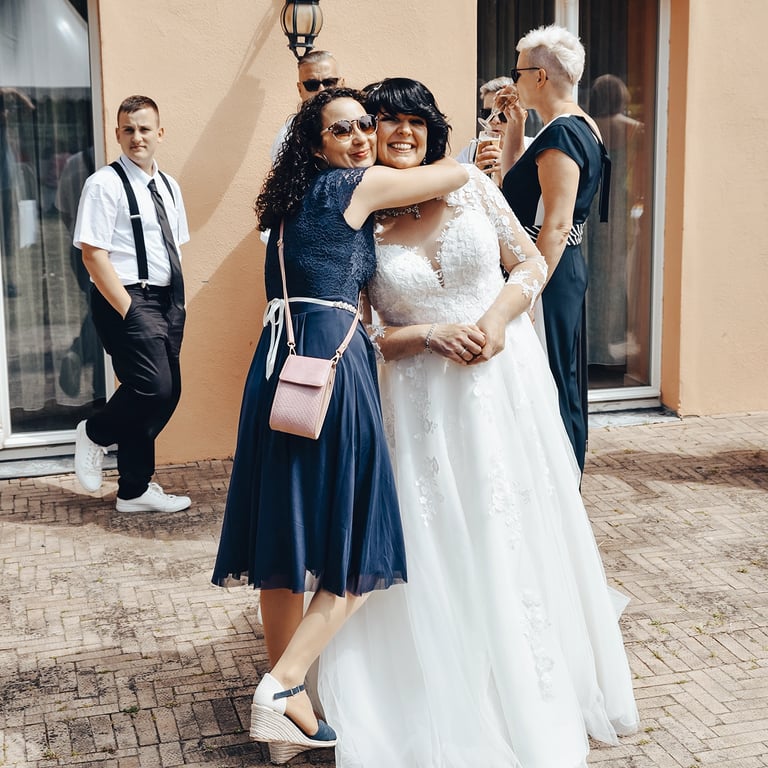 Eine Braut in einem weißen Hochzeitskleid wird von einer Freundin in einem dunkelblauen Kleid umarmt, während andere Hochzeit