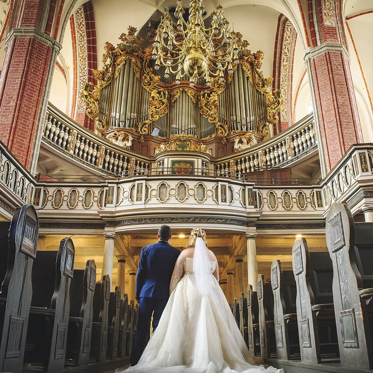  Ein Rückansicht-Foto eines Brautpaares, das in einer Kirche steht und auf eine große, verzierte Orgel blickt. Der Bräutigam 