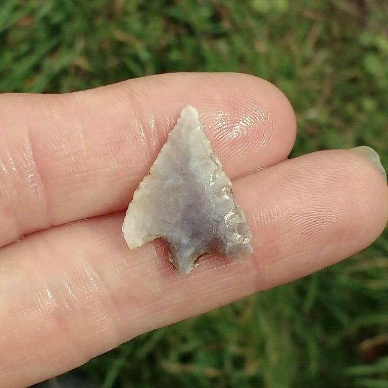 A tiny arrow shaped flint weapon, light grey, held on two fingers.