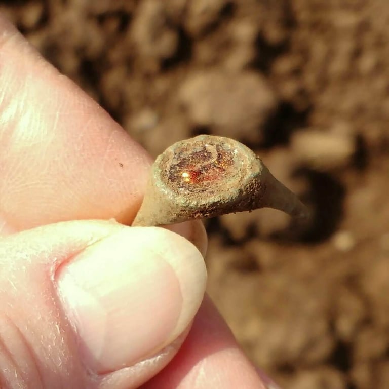 A broken bronze finger ring with green patina and orange inset stone, held between finger and thumb.