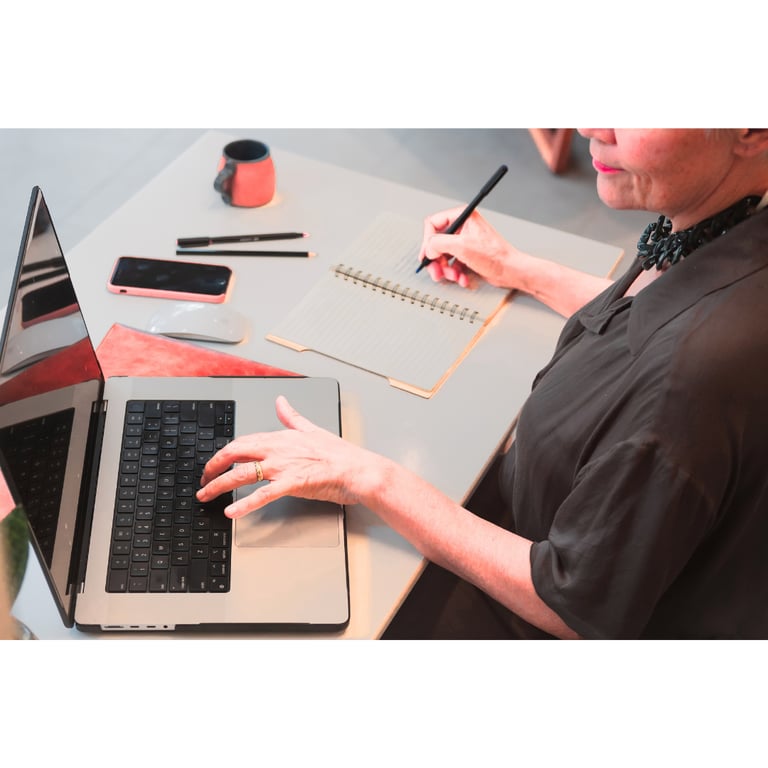 Uma mulher consulta o notebook aberto em cima da mesa enquanto faz anotações no caderno ao lado.