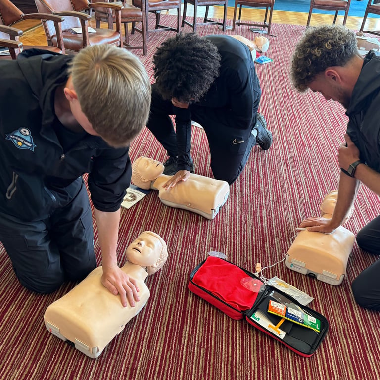 3 people kneel on the floor practicing CPR on adult training manikins, with an AED
