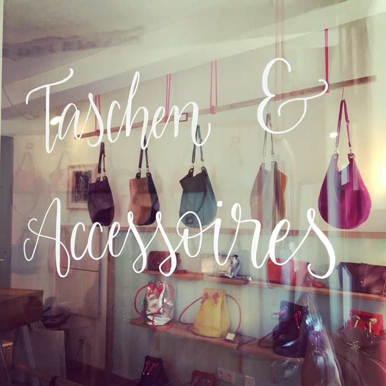 A stylish boutique window display featuring handbags and accessories with cursive white lettering.
