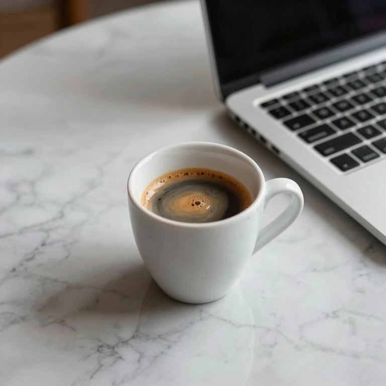 Close up of a cup of espresso on a clean marble desk next to a sleek laptop.