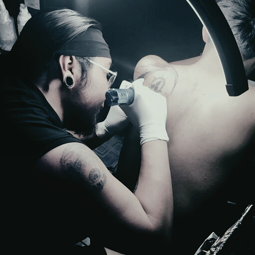 Tatuador profesional con guantes blancos aplicando tinta en el hombro de un cliente en un estudio.