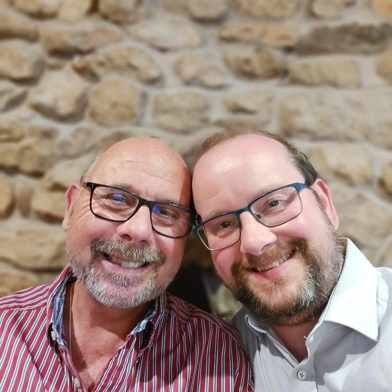 Two smiling men with glasses and beards pose together against a rustic stone wall background.