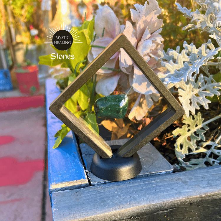 Polished green malachite crystal displayed in a black floating frame among garden flowers.