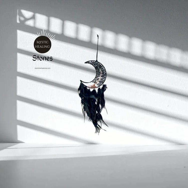 Obsidian crescent moon dreamcatcher with feathers hanging against a wall with window shadows.