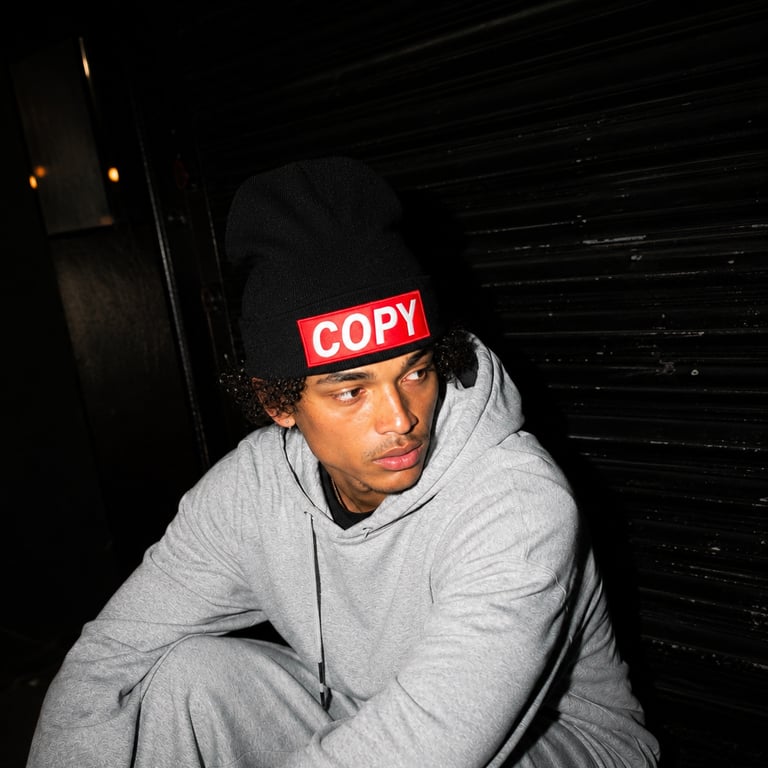 A man in a grey hoodie and black COPY logo beanie sits against a dark shutter door.