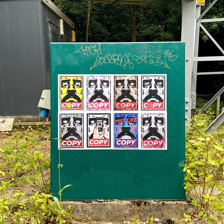 Urban street art stickers and graffiti tags on a green utility box in a park setting.