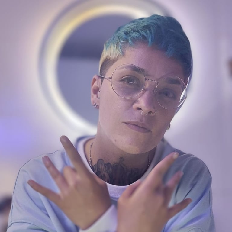 Persona con cabello corto azul y gafas, haciendo gestos con las manos frente a un aro de luz.