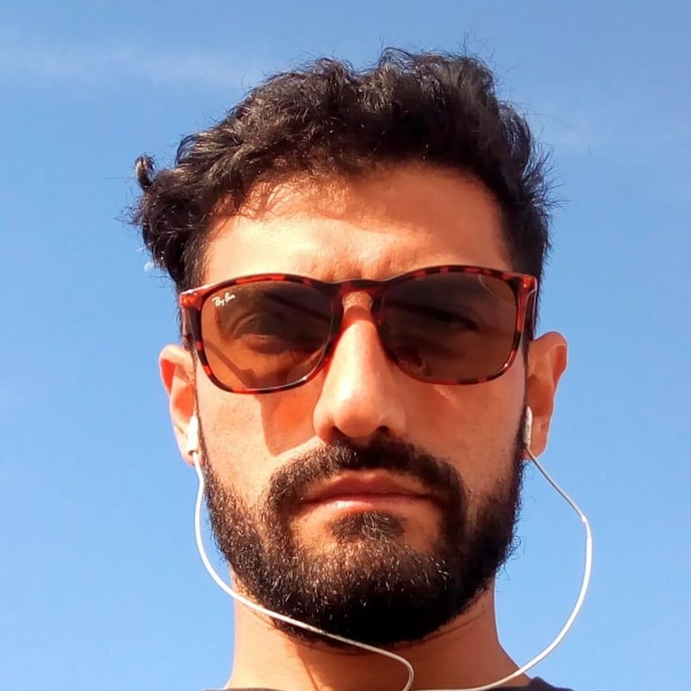 Hombre con barba usando gafas Ray-Ban de carey y audífonos blancos con cable, con el cielo azul 