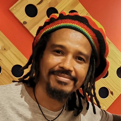 Un hombre con rastas, usando un gorro rasta colorido y una camisa gris, frente a una pared roja.