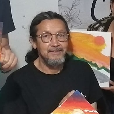 Un artista sonriente con gafas y perilla sosteniendo pinturas coloridas de paisajes al atardecer.