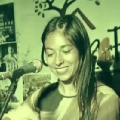 Fotografía vintage con tono verdoso de una cantante de folk sonriente, interpretando con micrófono
