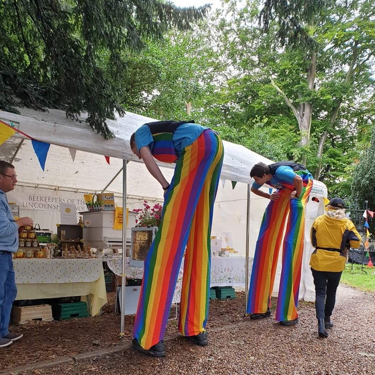 Two stilt walkers in rainbow striped pants visit a honey vendor stall at an outdoor festival.