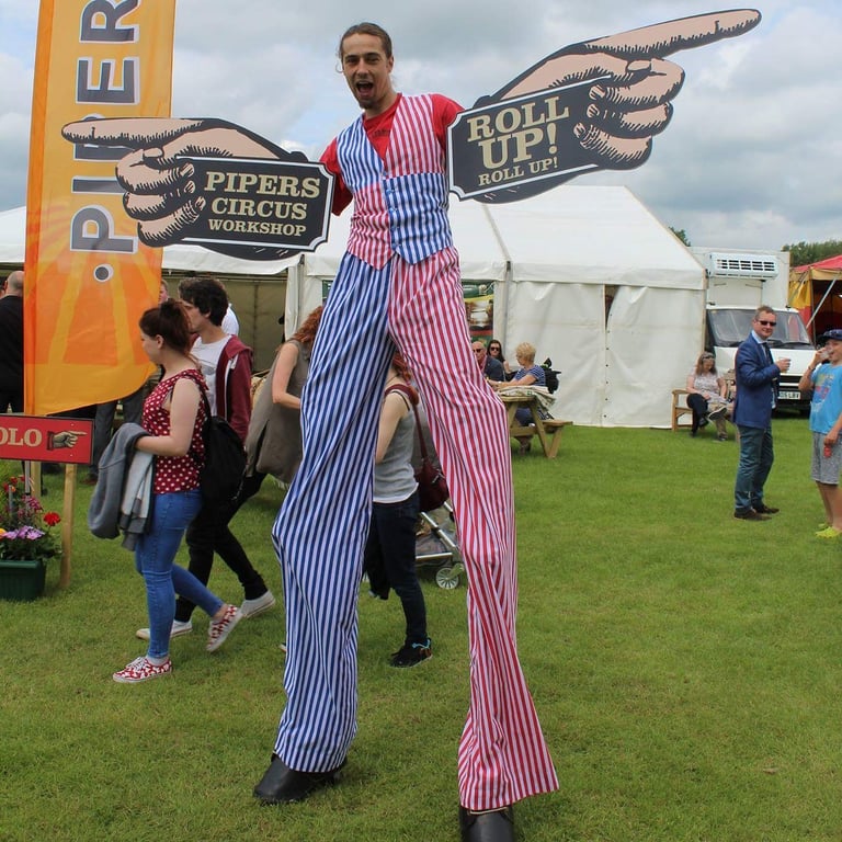 A circus stilt walker in red, white, and blue striped pants holds Pipers Circus Workshop signs at an outdoor festival.
