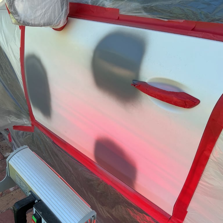 A white car door undergoing professional auto body paint repair with grey primer spots and red masking tape.