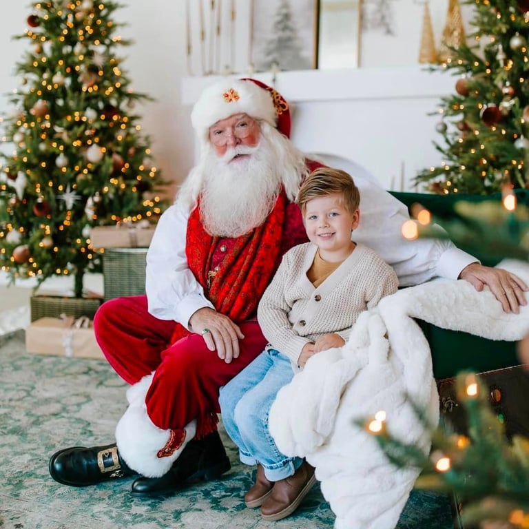 A smiling young boy sits with Santa Claus in a festive room with decorated Christmas trees.
