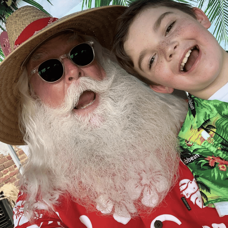 A smiling boy taking a selfie with Santa Claus wearing a summer straw hat and Hawaiian shirt.