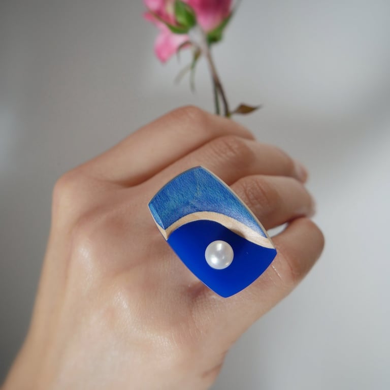 Hand wearing a blue statement wooden ring with a white pearl accent while holding a pink rose.