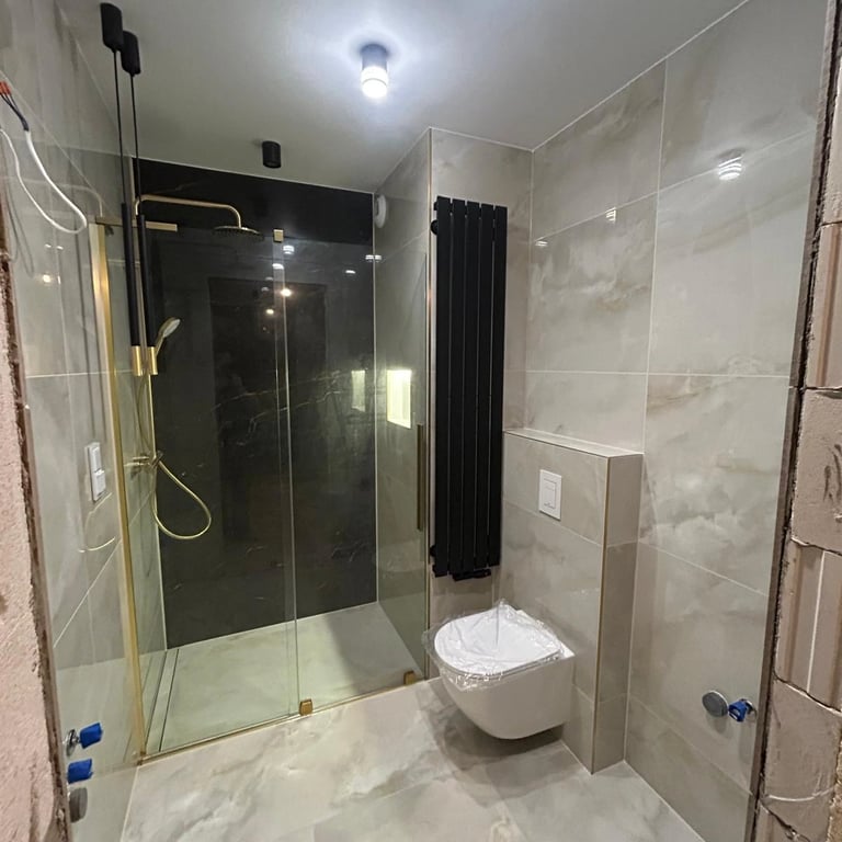 Łazienka z prysznicem w złotej ramie, marmurowe płytki i czarny grzejnik dekoracyjny.