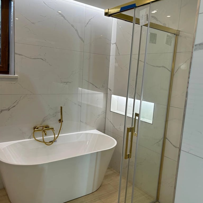 Łazienka z wanną i prysznicem w złotej ramie, marmurowe płytki i jasne oświetlenie.
